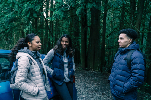 three people wearing winter jackets talking somewhere that looks like a forest or something near a blue car